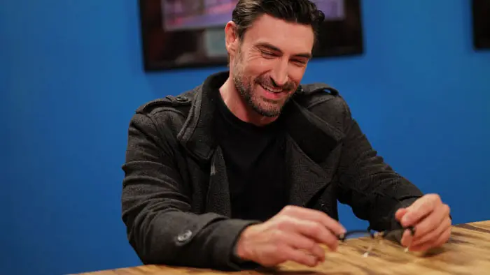 Ty Smith (John Wells) smiles at a table while holding his glasses in Learning You.
