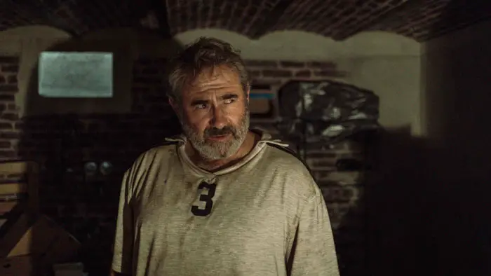 A bearded man in a dim basement looks off to the side in a tense moment in Maldoror (2026).
