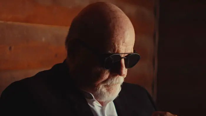 Michael Ironside as Dr. Jonathan Myers sits inside a cabin wearing dark sunglasses in warm, low light.