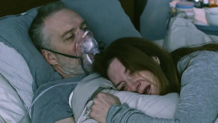 A woman clings to her incapacitated husband in bed as he wears an oxygen mask in Our Happy Place (2025).
