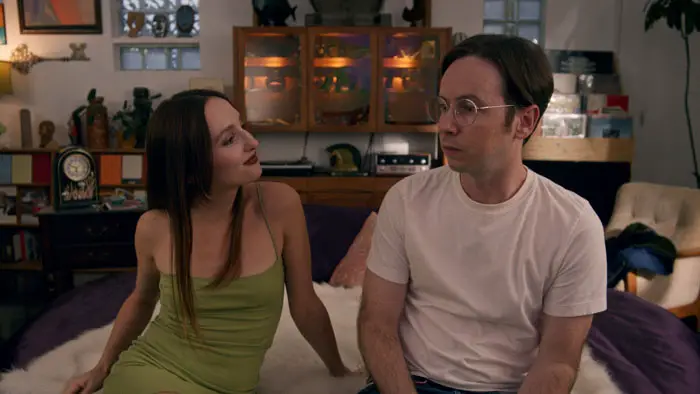 A woman and man sit on a bed talking in a cozy apartment in Paying for It (2026).
