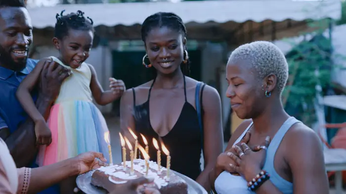 A group gathers outdoors around a birthday cake with lit candles, sharing a warm moment.