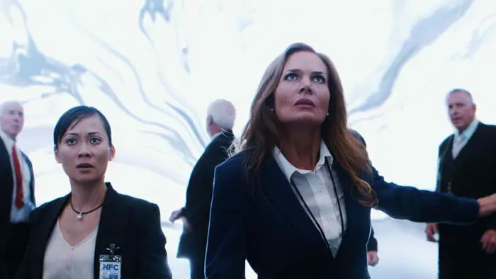 Two women with NFC badges stare upward as a bright portal-like phenomenon opens behind them in Space/Time (2026). 