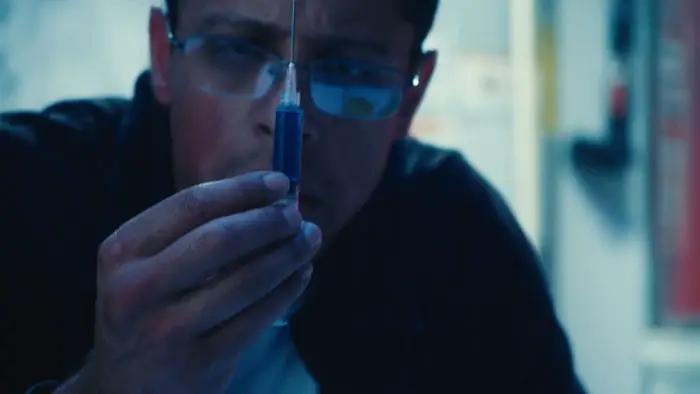 Richard holds a syringe of blue serum in The Geneticist (2026).
