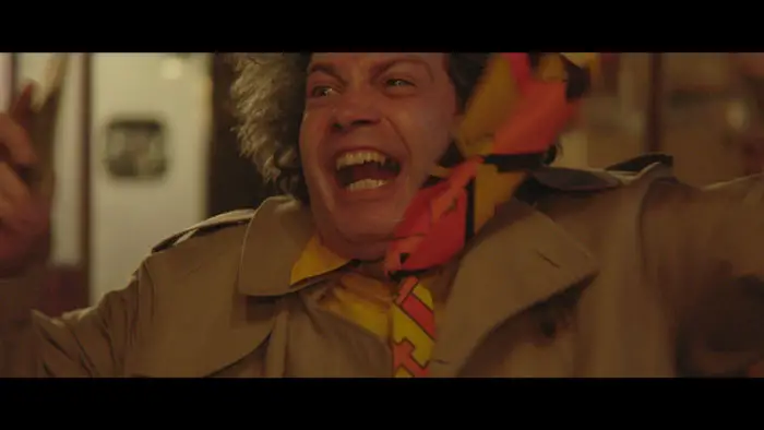 Brian (Brian Belott) in a trench coat and colorful tie, mid-outburst in The Man Without Qualities (2025). 
