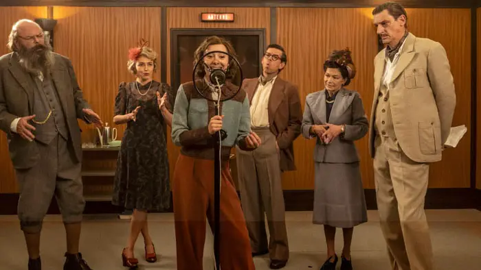 Scene from The Soundman (2025) with a performer at a vintage radio microphone as the studio ensemble looks on. 