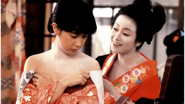 A geisha in a red kimono smiles while adjusting the robe of a young woman in Tokyo Bordello (1987).
