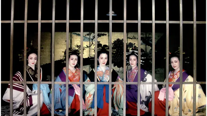 Five women in ornate kimono stand behind wooden bars in a Yoshiwara brothel interior in Tokyo Bordello (1987).