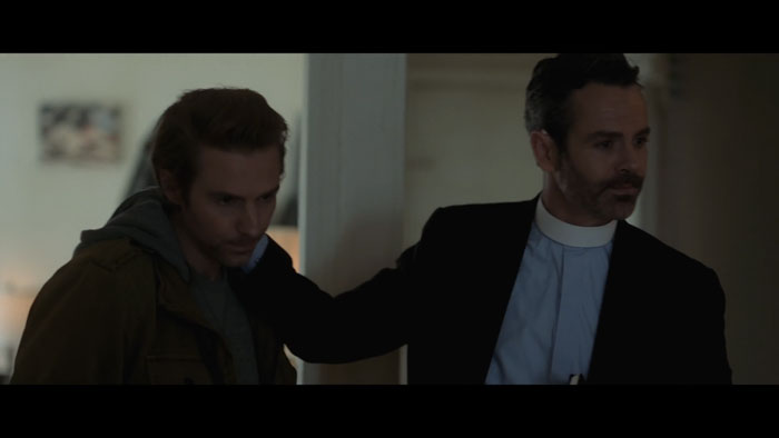 Two brothers enter a home in All Saints Day, with one wearing a priest collar and guiding the other inside.