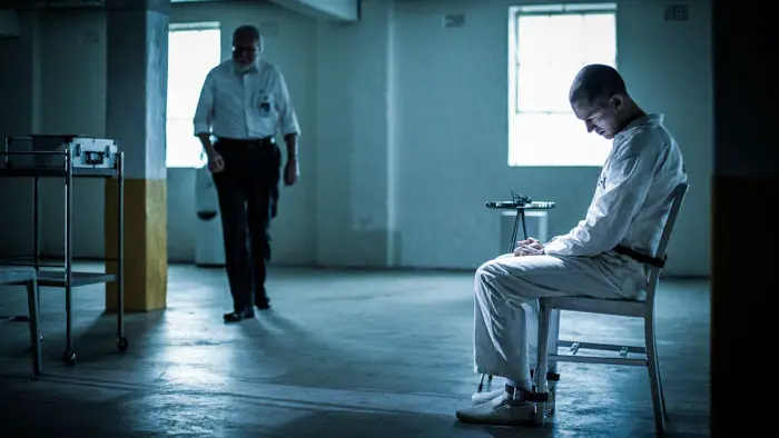 Prisoner sits in a chair under cold blue light as a guard approaches in Animus (2026).
