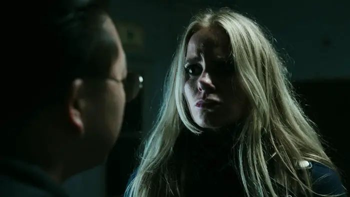 Tense close-up of a blonde woman confronting a man in a dimly lit scene from Last Hit (2026). 