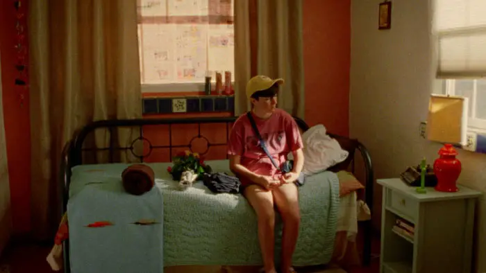 Nicole sits on a bed in a sunlit bedroom in Marlene Emilia Rios’ short film Lucia & Nicole.