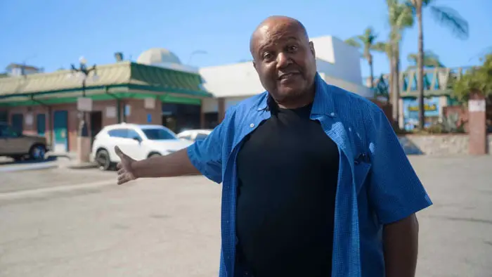 A man in a blue shirt gestures toward a storefront while speaking on camera in The Untold Story of No Retreat No Surrender (2026).