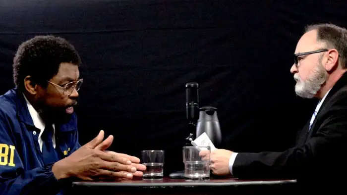 FBI Agent Justice Brown sits across from a podcast host discussing his paranormal ability to hear the dead in The Dead Guy (2026).